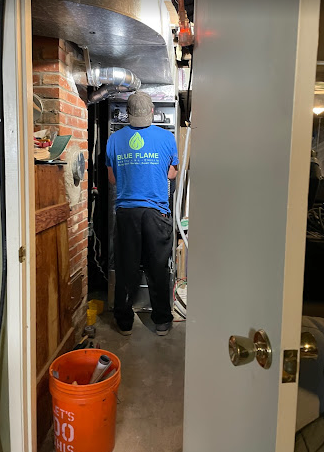 An HVAC technician in a blue "Blue Flame" shirt working on a Furnace inside a basement,