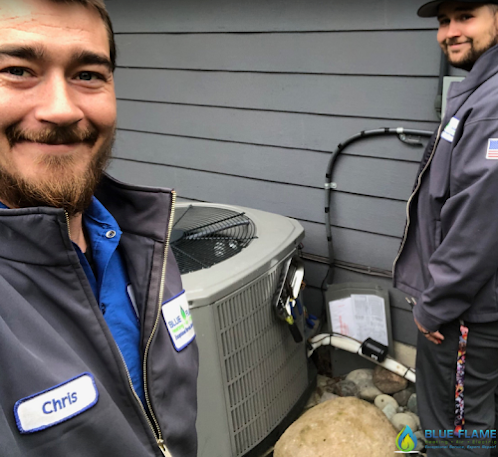 Two HVAC technicians from Blue Flame, wearing jackets with name tags, smiling and posing next to an air conditioning unit they just repaired outside a building.
