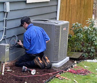 A Blue Flame HVAC technician wearing a blue shirt and black pants kneeling next to an air conditioning unit outside, working on the system's components.